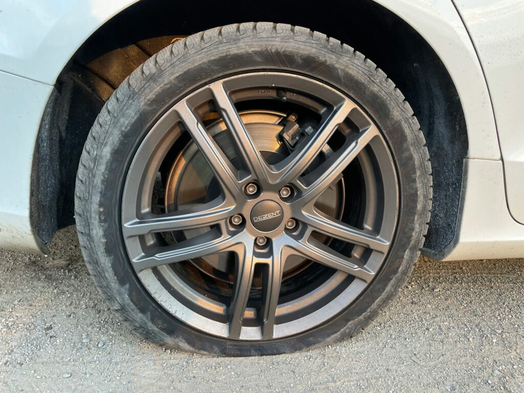 Side view of a flat tire mounted on a dark gunmetal Dezent split-spoke alloy wheel on a white car.