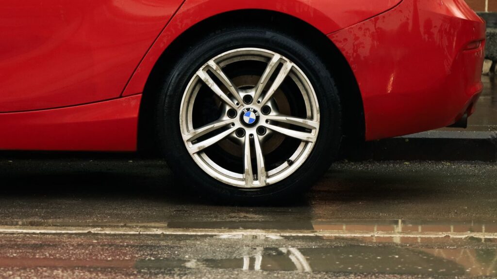 Side view of a silver double-spoke alloy wheel on a red BMW car parked on a wet street.