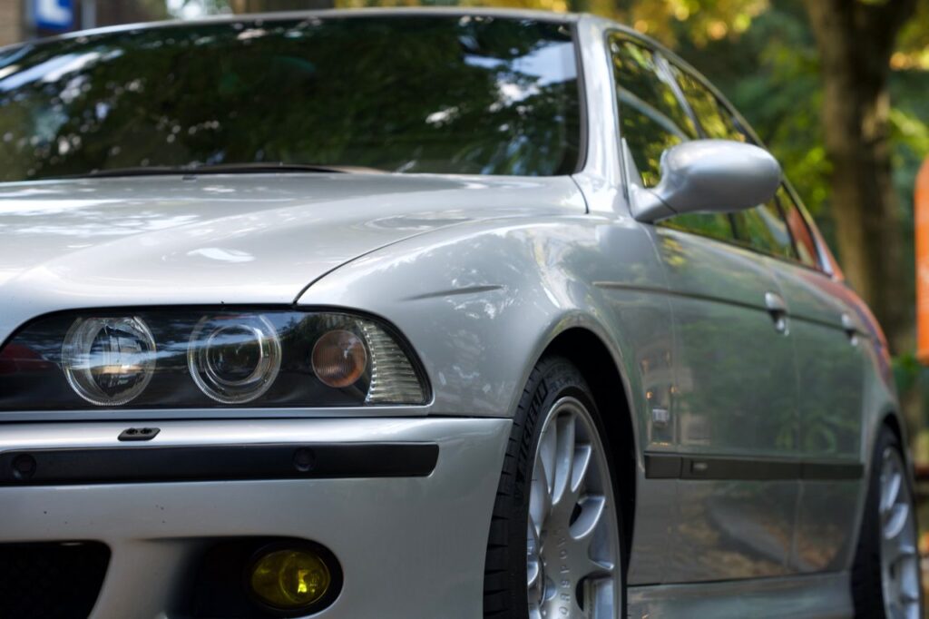 Low-angle front quarter view of a silver classic BMW E39 sedan showing angel eye headlights and yellow fog lights.